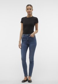 Černé přiléhavé tričko, modré skinny džíny s vysokým pasem, obuté s černými baletkami. Hladké materiály, minimální detaily, klasická silueta.