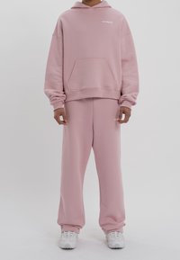 Personne portant un sweat à capuche oversize rose pâle et un pantalon de survêtement assorti avec des baskets blanches sur un fond uni.