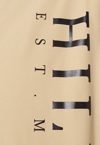 Beige stof med en glat tekstur, der viser store, dristige sorte bogstaver med teksten "EST." og et stiliseret "H"-design.