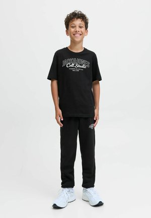 T-shirt en coton noir avec logo blanc, associé à un pantalon de survêtement noir comportant un petit logo. Les chaussures sont des baskets blanches.