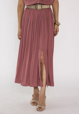 Femme portant une jupe midi plissée rose poudré avec une fente à l'avant, ceinture dorée, bracelets et sandales dorées à talons hauts à brides.