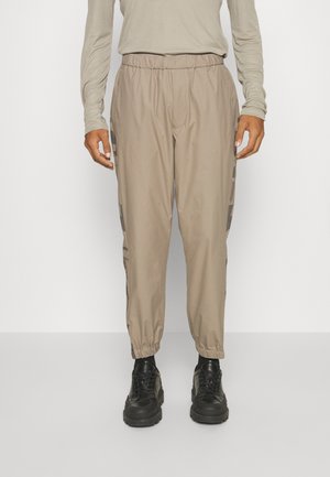 Pantalon beige à taille élastique avec détails graphiques sur les côtés, chevilles retroussées, porté avec des bottines noires épaisses à lacets et un haut beige clair à manches longues.
