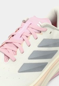 Άσπρο αθλητικό παπούτσι με ροζ λεπτομέρειες, με ανάγλυφο άνω μέρος, τρεις γκρι ρίγες και μαλακή σόλα. Τα κορδόνια είναι ανοιχτό ροζ.