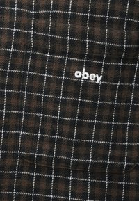 Tecido xadrez castanho e preto com linhas de grelha brancas. Apresenta o logótipo "obey" bordado em fio branco. Textura suave, design tecido.