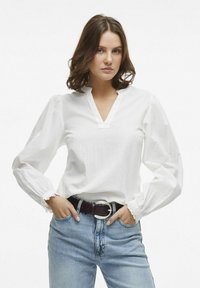 Hvid tekstureret bluse med V-udskæring, puffede ærmer og flæsemanchetter, parret med lyseblå jeans i denim og et mørkt bælte med en sølvspænde.