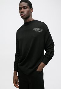 Pulover tricotat negru cu guler rotund ribat și mansete. Logo-ul brodat "JUST CAVALLI FIRED EARTH" pe piept. Textură netedă.