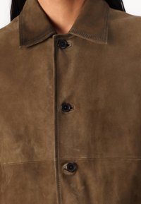 Palton din suede maro, cu nasturi, având un guler ascuțit, cu nasturi întunecați în contrast și o finisare moale, texturată.