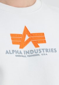 Hvid sweatshirt med et struktureret orange logo med stjernedesign og grå tekst "ALPHA INDUSTRIES, KNOXVILLE, TENNESSEE, U.S.A."