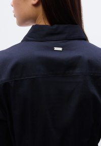 Πουκάμισο από ύφασμα σε ναυτικό μπλε χρώμα, με κλασικό γιακά και μικρή ασημένια πλακέτα με το λογότυπο στο πίσω μέρος, με λεία, ματ υφή.