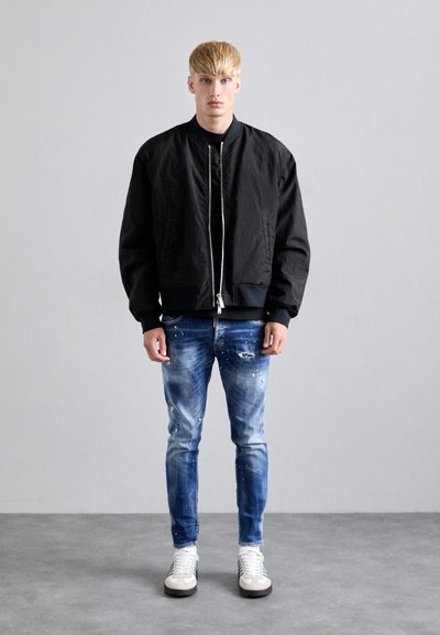 Jonge man met blond haar die een zwarte bomberjack, versleten blauwe spijkerbroeken en witte sneakers draagt terwijl hij tegen een grijze muur staat.
