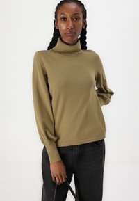 Pull en col roulé vert olive, texture douce, manches ballon longues, poignets côtelés, porté avec un jean noir, design minimaliste, coupe décontractée.