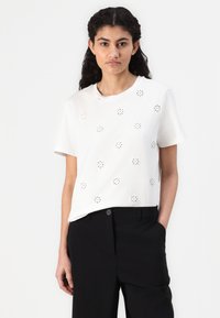 T-shirt en coton blanc à manches courtes avec des motifs floraux découpés. Associé à un pantalon noir, présenté sur un fond neutre.