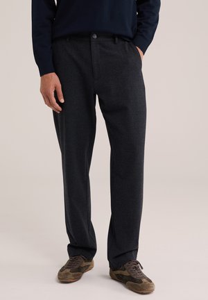Homme portant un pantalon gris foncé sur mesure et des baskets en daim marron, debout avec une main dans la poche sur un fond neutre.