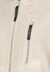 Fleece-Reißverschlussjacke in Cremefarbe, mit zwei Vorderreißverschlusstaschen mit kontrastierenden schwarzen Zippern und dezenter Logo-Stickerei am Saum.