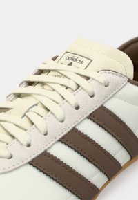 Άσπρο δερμάτινο αθλητικό παπούτσι με καφέ σουέντ λεπτομέρειες, τρεις καφέ ρίγες, στρογγυλά κορδόνια και ανάγλυφο λογότυπο στη γλώσσα.