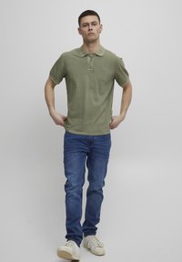 Olivnozeleni bombažni polo srajca s ploskim ovratnikom in dvema gumboma, v kombinaciji s slim-fit modrimi kavbojkami in belimi športnimi copati. Gladka tekstura, sproščen design.