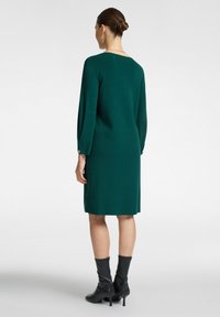 Grünes Strickkleid mit geradem Schnitt, langen Ärmeln und rundem Ausschnitt. Getragen mit schwarzen Ankle-Boots mit schmalem Absatz.