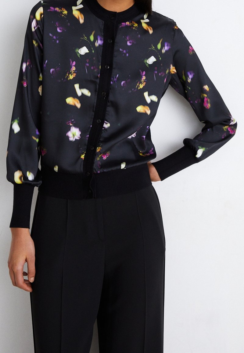 Chemisier noir à manches longues avec boutons et motif floral coloré, associé à un pantalon noir taille haute, main du modèle sur la hanche.