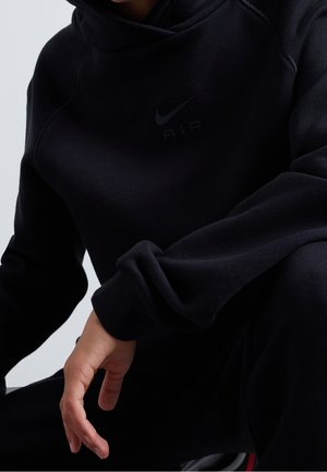 Osoba ubrana w czarną bluzę z kapturem Nike Air, lewa ręka widoczna spoczywa na zgiętym kolanie na neutralnym tle.