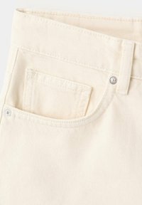 Gros plan sur un jean en denim couleur crème montrant la poche avant, la petite poche, les passants de ceinture et les rivets argentés sur la texture du tissu.