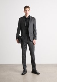 Donkergrijs op maat gemaakt pak met een textuurafwerking, gecombineerd met een zwart shirt en zwarte leren schoenen. Model houdt een zwarte clutch in de hand.