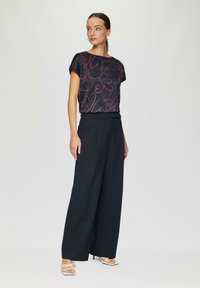 Schwarzes Oberteil mit rotem Herzmuster, kombiniert mit locker sitzenden, navyblauen Hosen. Das Modell trägt weiße Sandalen mit Riemchen. Neutraler Hintergrund.