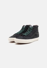 Čierne semišové sneakers so zvýšeným vrchom, zelenými šnúrkami, bielou gumovou podrážkou a polstrovaným golierom; obsahuje textúrované panely a logo značky na jazyku.