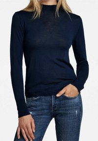 Frau in einem enganliegenden navyblauen Langarmtop und blauen Jeans, mit einer Hand in der Tasche vor einem weißen Hintergrund.
