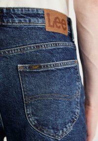 Σκούρα μπλε τζιν παντελόνι με καφέ δερμάτινη ετικέτα με το λογότυπο "Lee", αντίθετη ραφή και μια μόνη πίσω τσέπη με ετικέτα της μάρκας.