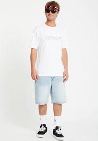 T-shirt blanche avec logo "Volcom", shorts en jean clair, baskets noires et lunettes de soleil rouges surdimensionnées. Design de tenue d'été décontractée.