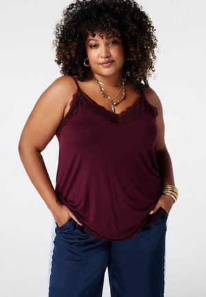 Zelfverzekerde vrouw met krullend haar draagt een bordeaux top met kanten randje, blauwe broek met witte strepen en gelaagde gouden sieraden, handen in de zakken.