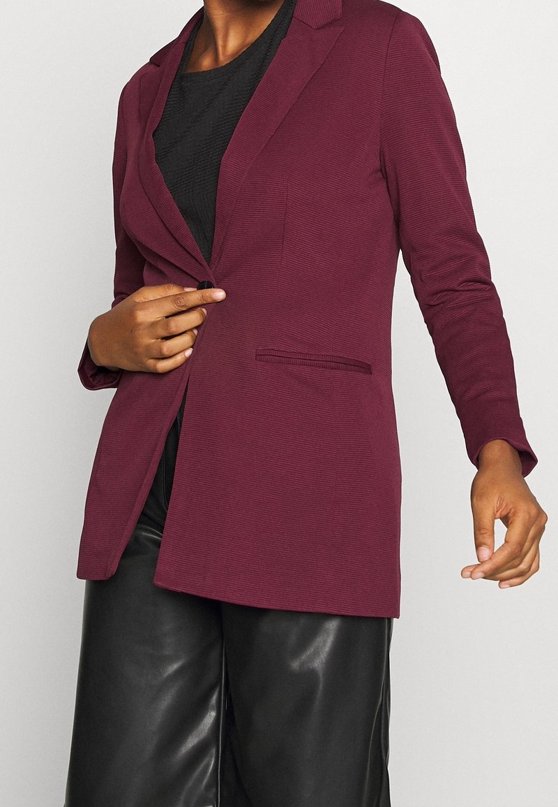 Personne portant un blazer rouge foncé sur un top noir et un pantalon en cuir noir, fermant le blazer d'une main.