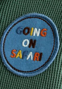 Grønt stof har en rund blå lap med den broderede tekst "GOING ON SAFARI" i forskellige farver, kantet med brun syning.