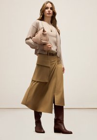 Gilet beige, jupe longue marron ceinturée avec fente sur le côté et poches, bottes marron jusqu'au genou, et un sac à main rose pâle.