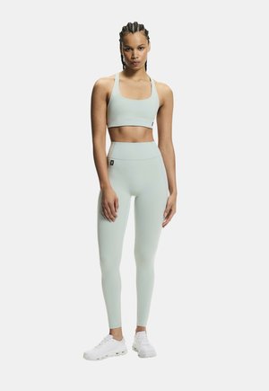 Vrouw staande in een lichtgroene sportbeha en legging met witte sneakers tegen een effen witte achtergrond.