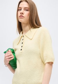 Νεαρή γυναίκα που φοράει ένα κρεμ πουλόβερ με κοντά μανίκια, μαύρα κουμπιά και γιακά, κρατώντας μία μικρή πράσινη τσάντα με ρυτίδες στο ένα χέρι.