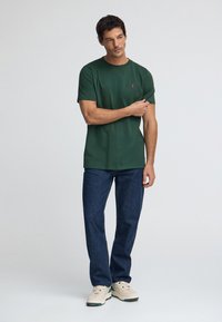 Grön bomulls-t-shirt med rund halsringning och korta ärmar, med en liten röd logotyp på bröstet; kombinerad med mörkblå denimjeans och ljusa sneakers.