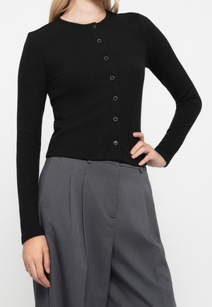 Femme portant un cardigan côtelé noir boutonné et un pantalon large plissé gris taille haute, posant avec une main sur la hanche.