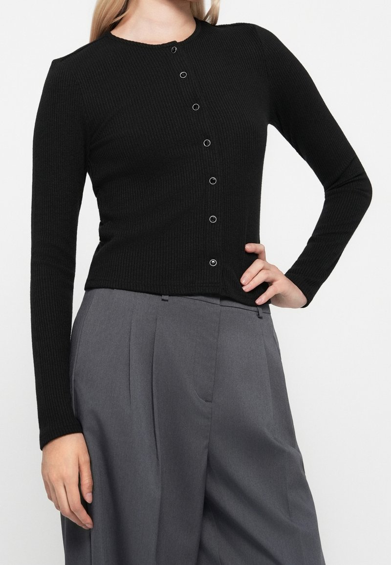 Femme portant un cardigan côtelé noir boutonné et un pantalon large plissé gris taille haute, posant avec une main sur la hanche.