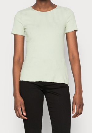 Osoba ubrana w jasnooliwkowy prążkowany top z krótkim rękawem i falowanym dołem, w połączeniu z czarnymi spodniami, na jednolitym tle.