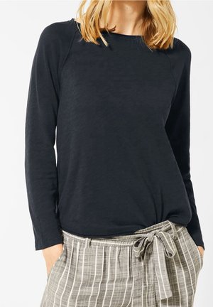 Kobieta ubrana w czarną bluzkę z długim rękawem wpuszczoną w beżowo-białe paskowane spodnie z wiązanym w pasie, ręce w kieszeniach.