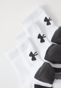 Calcetines deportivos blancos con textura acanalada, con acentos de logo negro y un diseño de lengüeta negra en el talón. Tres pares apilados.