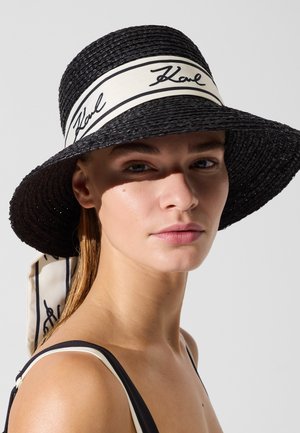 Jeune femme portant un chapeau noir tressé à large bord avec une bande blanche et une écriture noire signature, associé à un haut noir et blanc.