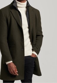 Manteau en laine olive avec un col à revers, doublure noir matelassée, trois boutons et poches latérales, porté sur un pull col roulé crémeux et un pantalon noir.
