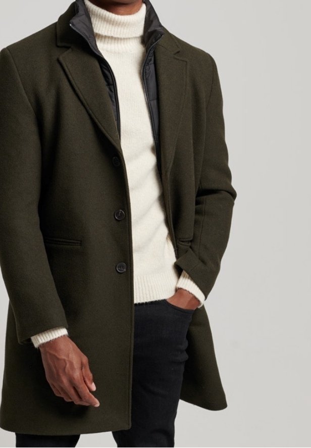 Manteau en laine olive avec un col à revers, doublure noir matelassée, trois boutons et poches latérales, porté sur un pull col roulé crémeux et un pantalon noir.