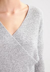 Gros plan sur une personne portant un pull en tricot gris clair texturé avec un col en V profond et un design croisé à l'avant.