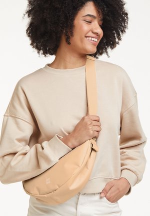 Lachende vrouw met krullend haar draagt een beige sweatshirt en witte broek, terwijl ze een tan crossbodytas met brede band over haar borst vasthoudt.