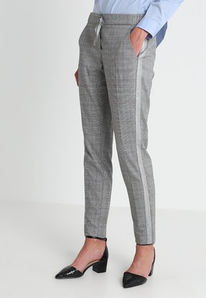 Frau trägt graue karierte, schmal zulaufende Hose mit Seitenstreifen und schwarze, flache Schuhe, steht vor einem schlichten hellen Hintergrund.