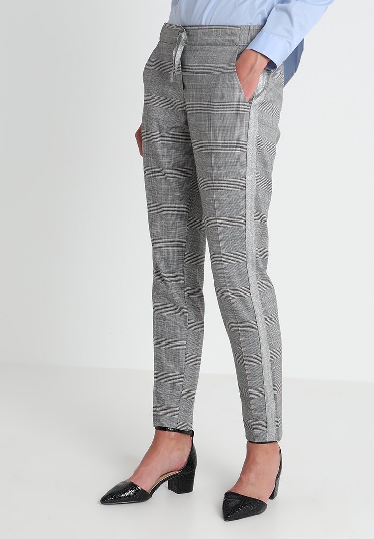 Femme portant un pantalon fuselé à carreaux gris avec des bandes latérales et des chaussures noires à talons bas, debout devant un fond clair uni.