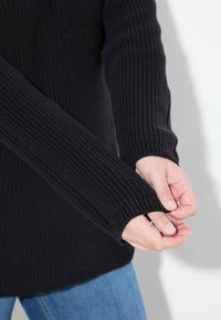 Maglione nero lavorato a maglia con texture a costine e maniche lunghe, che mostra una vestibilità ampia e un orlo arrotondato. Mani unite sulle maniche.